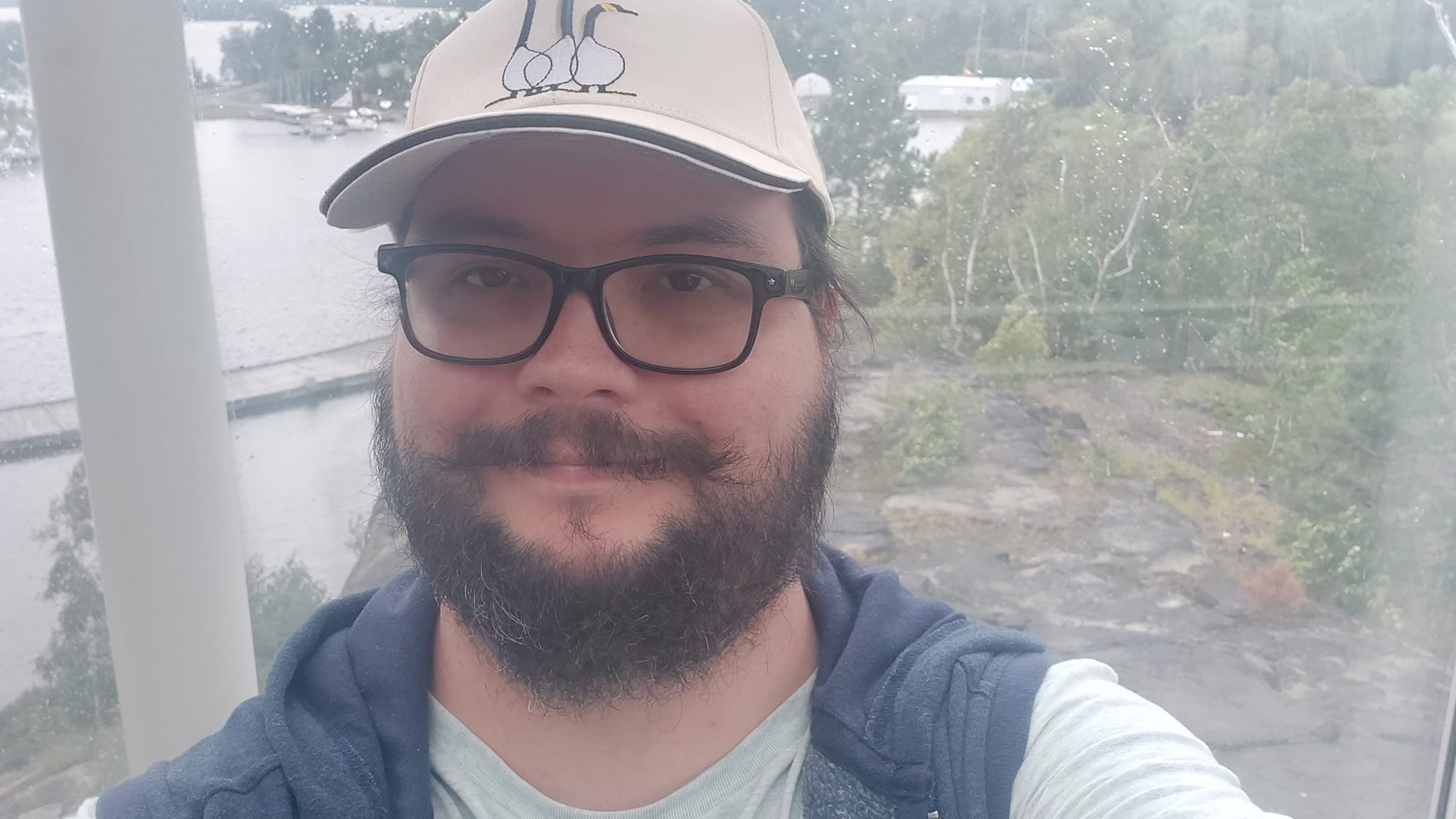 A man with a ball cap and bushy facial hair is standing in front of a window with a bedrock outcrop and lake visible in it